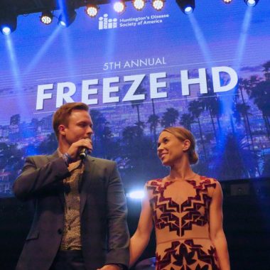 Scott and Kelsey Porter hold hands and stand on stage at the 2019 Freeze HD event. Scott holds a microphone and looks into Kelsey's eyes.