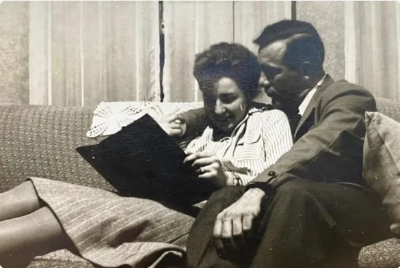 In an older, black and white photo, a woman reclines against a man on a sofa. She's in a dress and he's in a suit, and both are looking at what appears to be a book.