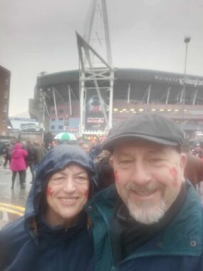 A cloudy and rainy photo shows a smiling couple in their 60s standing outside in front of a large football or rugby stadium. It's raining and they're both wearing jackets. The woman has a blue hood pulled up and the man is wearing a cap. A large crowd mills about in the background, just in front of the stadium. 