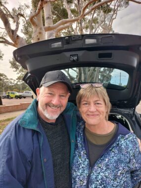 A couple - a man on the left and a woman on the right - in their 60s pose with broad smiles in front of the raised back door of an SUV or other type of vehicle. It looks like they're in an outdoor parking lot. They're both wearing light jackets and the day seems overcast.