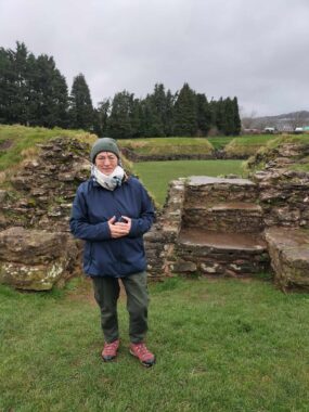 A woman wearing a rain jacket, scarf, and warm cap stands on the grassy lawn before ancient Roman ruins, with fir trees (or similar) in the background. The sky above is gray and cloudy and there isn't much light. It looks like it's been raining. 