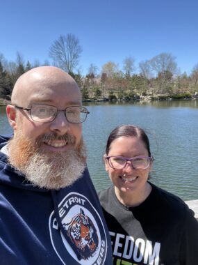 A man and woman smile for a photo while standing in front of a lake on a sunny day.