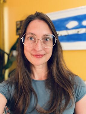 A headshot shows a woman with long brown hair and glasses standing in front of a yellow wall.