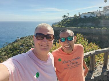 Two men, each wearing sunglasses and T-shirts, stand in front of a wooden midheight barrier with open slots. Behind them is what appears to be ocean and a couple of levels of land with plenty of flora.
