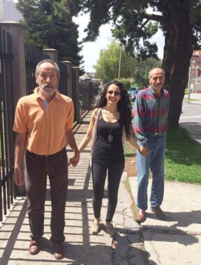 Two men and one woman - from the same family - hold hands as they walk down a sidewalk on a tree-lined street on a sunny day.