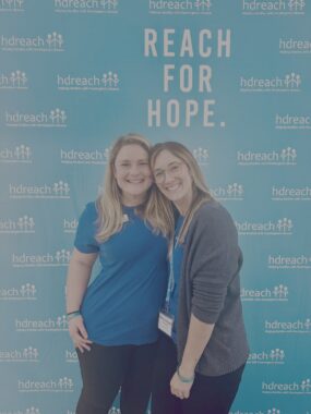 Two women, both wearing blue T-shirts, pose together for a photo in front of a backdrop that says "Reach for Hope" and has the HD Reach logo.