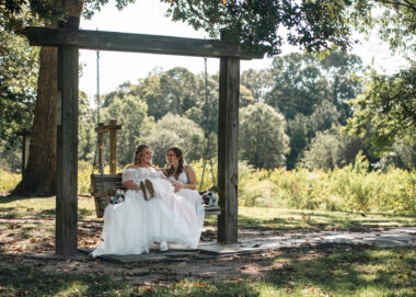 Two women, both wearing long white wedding dresses, sit together on an outdoor wooden swing. It's a sunny day but they are in the shade, surrounded by big trees.