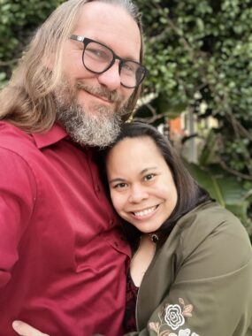 A man and a woman - husband and wife - smile for a close-up photo. They are standing facing each other and hugging, but their heads are turned toward the camera. There are trees and greenery in the background.