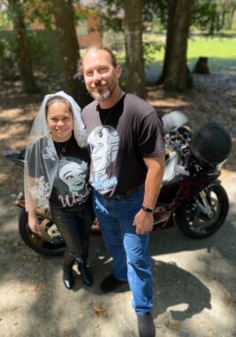 A couple - husband and wife - pose for a photo in front of a motorcycle. Both are in casual clothes but the woman is also wearing a bridal veil. It's a sunny day outside and they're smiling broadly.