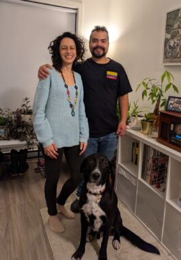 A husband and wife stand in their living room with a black-and-white dog sitting in front of them. The man has his arm around his wife and is wearing a T-shirt with the Colombian flag on it.