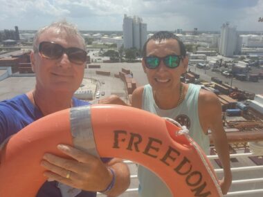 Two men stand with a background of what appears to be a terminal near a city. The man on the left, in a blue T-shirt and sunglasses, holds an orange life ring with the word "Freedom" on it. The man on the right wear sunglasses and a pale green tank top.