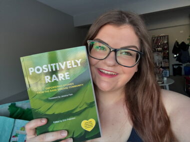 A woman in a selfie holds up a book titled "Positively Rare." She's in a living room. The smile on her face indicates that she's proud of the book.