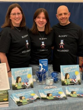 A teenage girl stands between her parents in front of a table displaying the children's book she wrote. All three are wearing matching black T-shirts that say "Bloopy has landed," in reference to a character in the book.