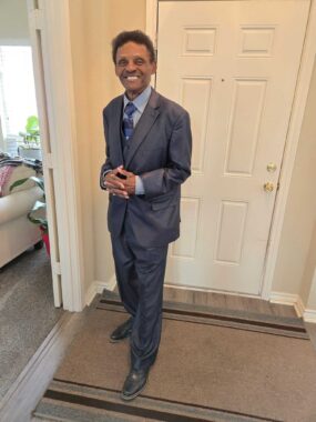A man is pictured wearing a suit and tie, and standing in what appears to be the foyer of his home. He's smiling and has his hands clasped in front of him.