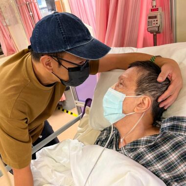 An older woman is lying in a hospital bed wearing a flannel gown and a face mask. Her adult son is standing beside the bed and leaning close to her, with his left hand gently cradling her head. He's wearing baseball cap and a face mask. Even though half of their faces are covered, the love and connection they share is evident.