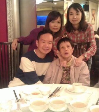 A family of four - a mom and three adult children - pose for a photo during a meal. The mom and her son are seated side by side in chairs, while the two daughters stand behind them. A round table in front of them is covered with bowls of food.