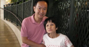 A middle-aged man smiles for a photo with his mother. They are seated in front of a railing in what appears to be a hallway.