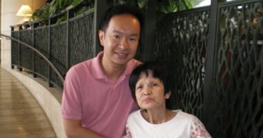 A middle-aged man smiles for a photo with his mother. They are seated in front of a railing in what appears to be a hallway.