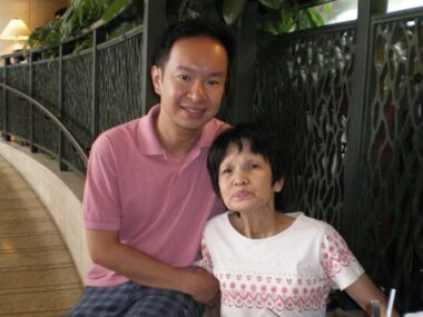 A middle-aged man smiles for a photo with his mother. They are seated in front of a railing in what appears to be a hallway.