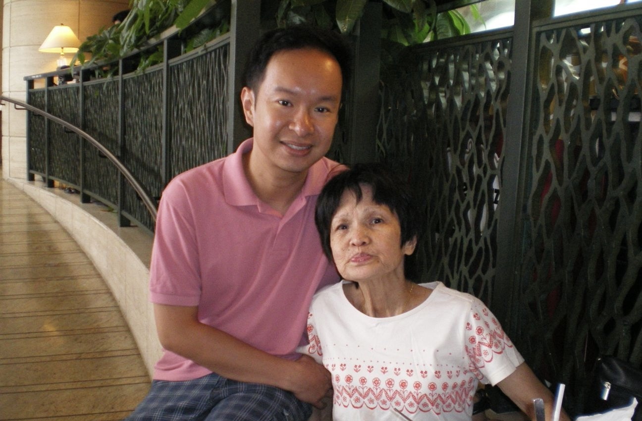 A middle-aged man smiles for a photo with his mother. They are seated in front of a railing in what appears to be a hallway.