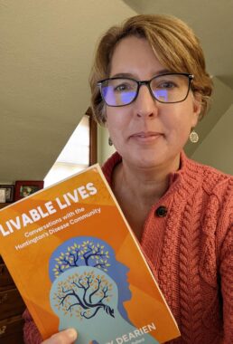 A woman smiles while holding up a copy of her book, "Livable Lives: Conversations with the Huntington's Disease Community." She's wearing glasses and a coral sweater and appears to be inside her home.