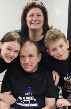 A family of four - husband, wife, son, and daughter) pose in a circle. Their black T-shirts read, "“All I Want for Christmas is Huntington’s cure.”