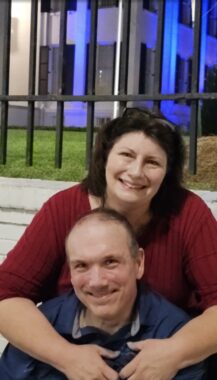 A husband and wife pose before a building lit with blue lights. The woman sits behind the man, with her arms around him. 