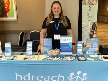 A woman stands behind a booth with HD Reach on it.