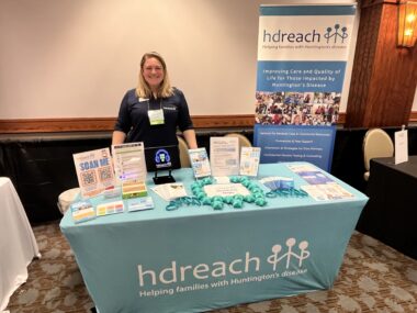 A woman stands at a table filled with Huntington's disease information.